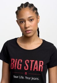 Juodas medvilninis marškinėlis su užrašu "BIG STAR" nublukintu raudonu šriftu, apvalia iškirpte ir trumpomis rankovėmis.