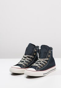 Baskets montantes en toile bleu foncé avec des lacets beiges, œillets dorés et embout en caoutchouc blanc. Présentent une semelle blanche profilée avec des accents rouges.