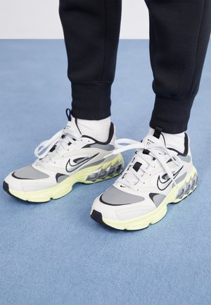 Baskets de sport blanches et grises avec semelles rembourrées jaunes, portées avec un pantalon noir et des chaussettes blanches sur un sol recouvert d’un tapis bleu.
