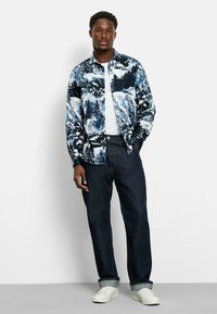 Kurzärmliges Hemd mit Knöpfen im blau-weißen Bergmuster, kombiniert mit dunklen Jeans und weißen Schuhen, bietet eine lässige Passform und einen entspannten Stil.