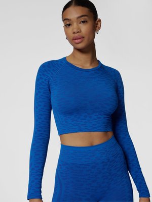 Kobieta w dopasowanym niebieskim crop topie z długim rękawem i dopasowanych legginsach z wysokim stanem z subtelnym wzorzystym wzorem.