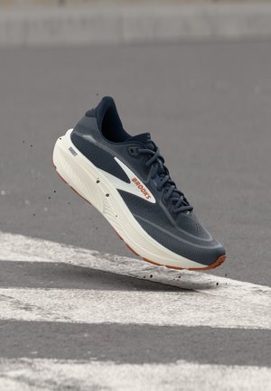 Scarpa da corsa Brooks blu scuro con dettagli bianchi e arancioni, a mezz'aria sopra asfalto con segnaletica stradale bianca e piccole particelle di ghiaia.