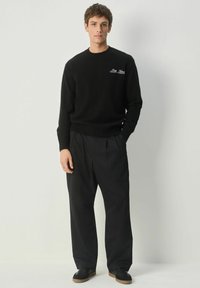 Ung mand står med den ene hånd i lommen iført en sort sweater med tekst, løse sorte bukser og sorte sko foran en ensfarvet baggrund.
