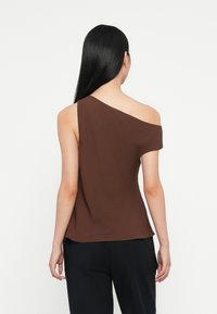 Top marrone a spalla unica realizzato in tessuto morbido con maniche corte. Taglio dritto, texture liscia e vestibilità rilassata. Indossato con pantaloni neri.