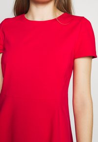 Nainen, jolla on yllään kirkkaan punainen lyhythihainen mekko, jossa on istuva vyötärö ja pyöreä pääntie, näkyy olkapäistä ylös rintakehään.