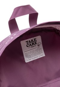 Interior de una bolsa de tela morada con un asa trenzada. Presenta una etiqueta blanca con el texto "CUIDA de PEANUTS". Textura suave, diseño minimalista.