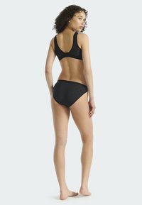 Žena s kudrnatými vlasy, oblečená v černé vzorované podprsence a odpovídajících vysokých kalhotkách, stojící bosky, otočená zády k jednoduchému pozadí.