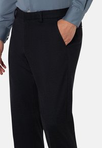 Schwarze Hose mit strukturiertem Gewebe. Verfügt über einen flachen Bund, seitliche Taschen und Gürtelschlaufen. Kombiniert mit einem hellblauen Hemd.