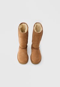 Paar mittelhohe Stiefel aus braunem Wildleder mit cremefarbenem Futter aus Fleece, aufrecht stehend vor weißem Hintergrund.