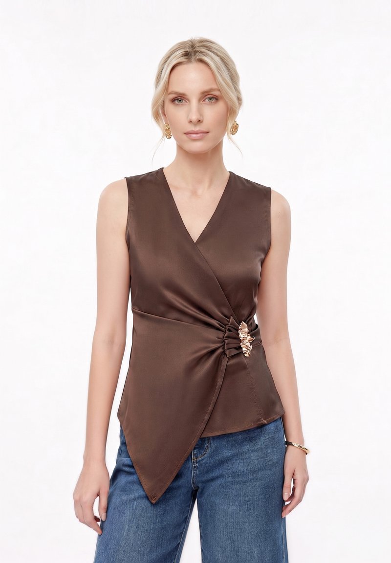 Femme blonde portant des boucles d’oreilles et un bracelet en or, un top marron sans manches drapé avec détail froncé, et un jean bleu sur fond blanc.