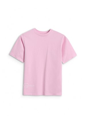 Dziewczyna o długich brązowych włosach nosi oversize różowy T-shirt na białej koszulce z długim rękawem, bordowe spodnie i czarne trampki, stojąc na tle białej ściany.