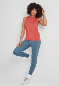 Woman in coral sleeveless top and blue leggings stands on one leg, touching curly hair, against plain light gray background.