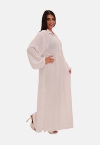 Longue robe blanche en tissu texturé avec un col, des boutons décoratifs et des manches volumineuses, présentant une coupe fluide et un design longueur sol.