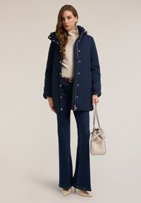 Chaqueta acolchada azul marino con cierre de cremallera y botones a presión, con capucha y herrajes dorados, combinada con suéter crema, jeans acampanados azules y bolso beige.