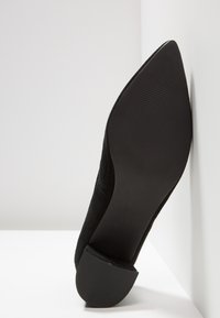 Chaussure noire à bout pointu avec semelle texturée et petit talon bloc, positionnée verticalement contre des surfaces blanches.