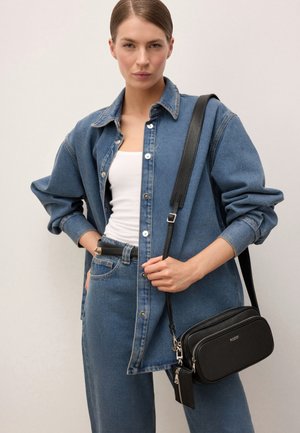 Žena v džínsovej bunde a džínsoch, bielom tielku, drží čiernu kabelku cez rameno s nastaviteľným popruhom na jednoduchom svetlom pozadí.