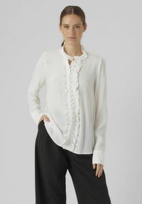 Witte blouse van lichtgewicht stof, met een gerimpelde halslijn en knoopsluiting aan de voorkant. Lange mouwen met manchet details.
