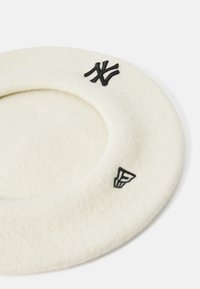 Biały filcowy beret z czarnymi haftowanymi logo z przodu. Okrągły kształt z miękką teksturą i niskim profilem.