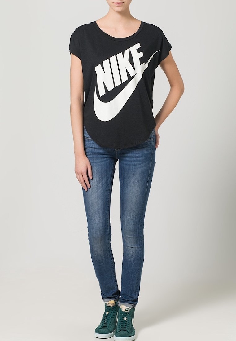 Zwarte T-shirt met korte mouwen met een groot wit Nike-logo, gecombineerd met blauwe skinny jeans en groene hoge sneakers. Effen achtergrond.