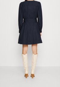 Vestito navy a maniche lunghe, vita aderente, gonna a goni e orlo a strati. Abbinato a stivali crema fino al ginocchio con tacco largo.