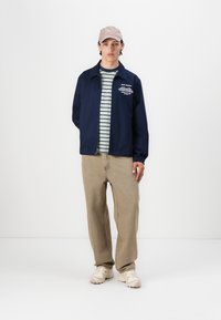 Navy zip-up jacket, striped shirt with green and white lines, beige baggy jeans, light-colored sneakers, and a beige cap.