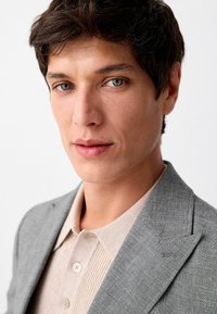 Szary żakiet wykonany z teksturowanej tkaniny, na beżowej dzianinowej koszulce z guzikami. Zbliżenie na rysy twarzy modelki i niebieskie oczy.