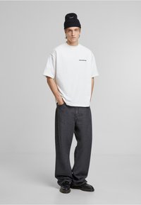 Weißes T-Shirt mit kurzen Ärmeln und dem Aufdruck "VERBOTEN", kombiniert mit lockeren, dunkelgrauen Jeans und schwarzen Schnürschuhen, ergänzt durch eine schwarze Strickmütze.