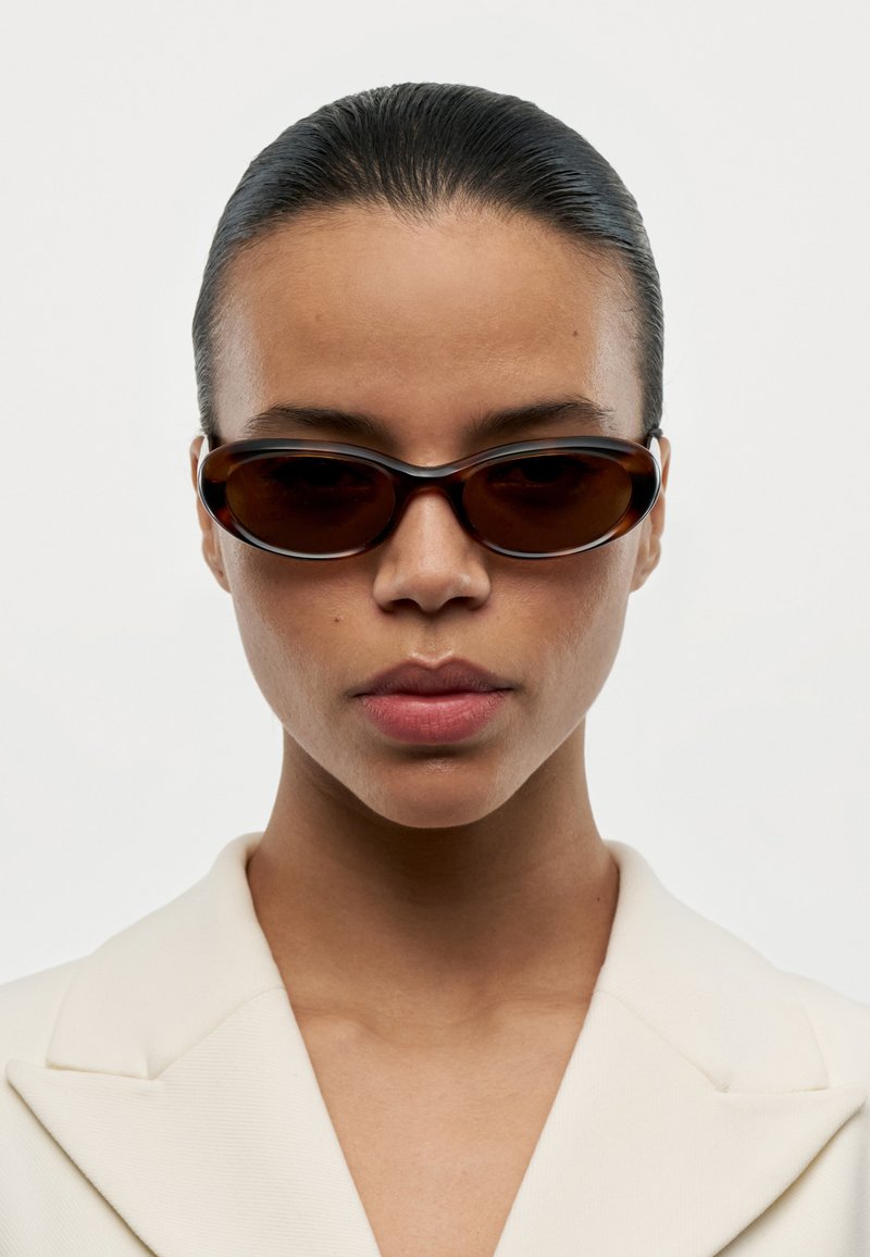 Mujer con cabello oscuro y liso, lleva gafas de sol estrechas y oscuras y un blazer crema, mirando hacia adelante contra un fondo claro y liso.