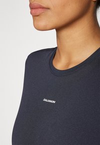 Tumed sinine aktiivne T-särk ümmarguse kaelusega, tekstuuriga kangas ja valge "SALOMON" logo rinnal. Lähedalne vaade kaelusele ja ülemisele kehale.