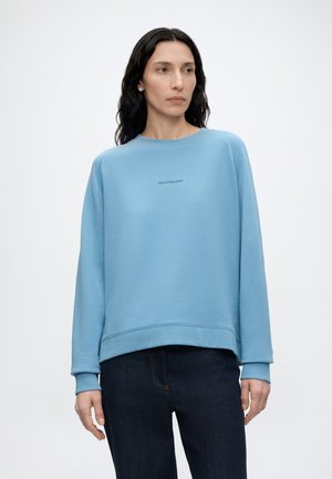 Kvinde med langt mørkt hår iført en lyseblå sweatshirt med lille tekst i midten og mørke jeans, stående mod en hvid baggrund.