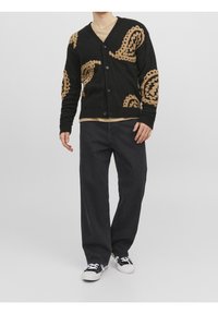 Cárdigan negro con un patrón de paisley en color marrón, con botones en la parte delantera, puños y dobladillo acanalados. Lleva una camisa beige y está combinado con pantalones negros.