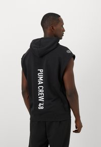 Man in een zwarte mouwloze hoodie met de tekst "PUMA CREW '48" verticaal op de rug, staand in profiel tegen een effen lichte achtergrond.