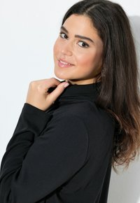 Mustavakuru turtleneck-neule, jossa on tiukat pitkät hihat. Malli poseeraa sivuprofiilissa, tuoden esiin sileän tekstuurin ja hienovaraisen kiillon.