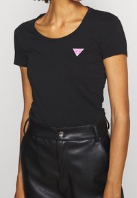Zwarte katoenen t-shirt met een ronde halslijn en korte mouwen, met een roze driehoeklogo op de linkerborst. Gepaars met zwarte leren broek.