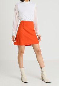 Falda roja acampanada hecha de tejido texturizado, combinada con una blusa blanca de cuello alto y botas tobilleras beige con tacón cuadrado y cremallera lateral.