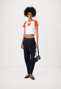 Cropped white t-shirt with orange sleeves and heart graphic, paired with dark blue jeans and black bag. Model wearing black flats.