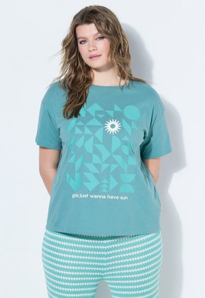 Kobieta o falowanych włosach nosi turkusowy t-shirt z geometrycznym wzorem słońca i napisem „girls just wanna have sun”, w połączeniu z turkusowo-białymi spodnimi wzorami.