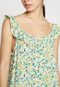 Femme portant une robe sans manches à fleurs avec un col à volants dans des tons verts, jaunes et blancs, montrée des épaules jusqu'au milieu du torse.