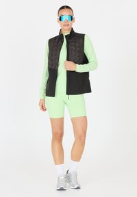 Gilet imbottito nero sopra una maglietta a maniche lunghe verde chiaro e pantaloni corti da bici abbinati. Sneakers argento e grandi occhiali da sole blu specchiati.