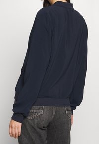 Marinblå bomberjacka med ribbad nederkant och ärmslut, med en slät yta och avslappnad passform, visad bakifrån.