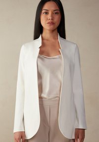 Weißer, langärmeliger Mantel mit Stehkragen, aus glattem Stoff. Trägt man über einem leichten Satin-Oberteil, kombiniert mit beigen Hosen.