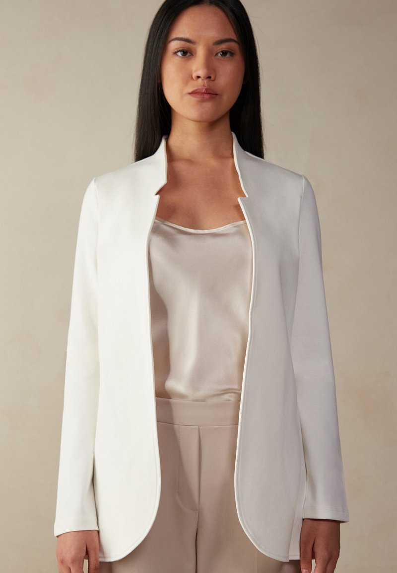 Chaqueta blanca de manga larga con cuello mandarín, hecha de una tela suave. Lleva una blusa de satén clara debajo, combinada con pantalones beige.