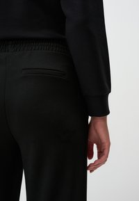 Czarne dresy o gładkiej fakturze, z elastycznym pasem i małą kieszonką z tyłu. Ręka umieszczona nieco na biodrze.