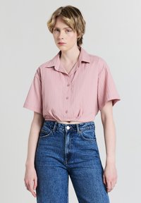 Camicetta corta a maniche corte di colore rosa chiaro con texture a righe verticali e chiusura con bottoni. Indossata con jeans in denim blu a vita alta.