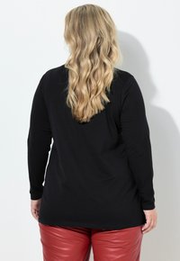 Osoba s dlouhými vlnitými blond vlasy, oblečená v černém topu s dlouhými rukávy a červených kožených kalhotách, čelící bílé zdi.