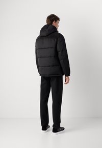 Casaco acolchoado preto com capuz, apresentando seções acolchoadas e punhos elásticos, combinado com calças pretas e sapatos pretos sem atacadores.