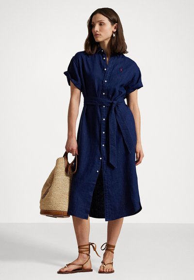 Jūras zila lina kleita ar īsām sarullētām piedurknēm, pogām pa visu priekšpusi un atbilstošu jostu. Valkāta ar sandālēm uz siksniņām un austotu somu.