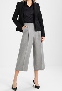 Culotte grigie in misto lana con vita alta, dotate di pieghe frontali, abbinate a una camicia nera con bottoni e a un blazer nero. Tacchi a punta neri.