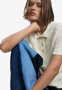 Polo shirt in maglina color crema con motivo floreale e tre bottoni; abbinato a jeans nelle sfumature di blu chiaro e scuro, con texture diverse.
