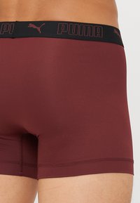 Boxeri din țesătură roșie, cu un elastic negru în talie, având logo-ul "PUMA" și o mică siluetă a unei pisici. Textură netedă, cu un design potrivit.